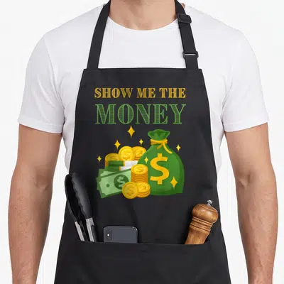 Wealthy money apron - Show me the money text above green money bag, gold coins, and banknotes with sparkles — shown on a black cotton canvas apron against a white background