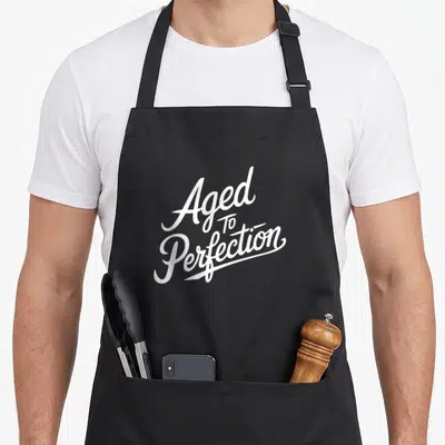 Aged perfectionist apron - White stacked script text reading Aged To Perfection on grey background — shown on a black cotton canvas apron against a white background
