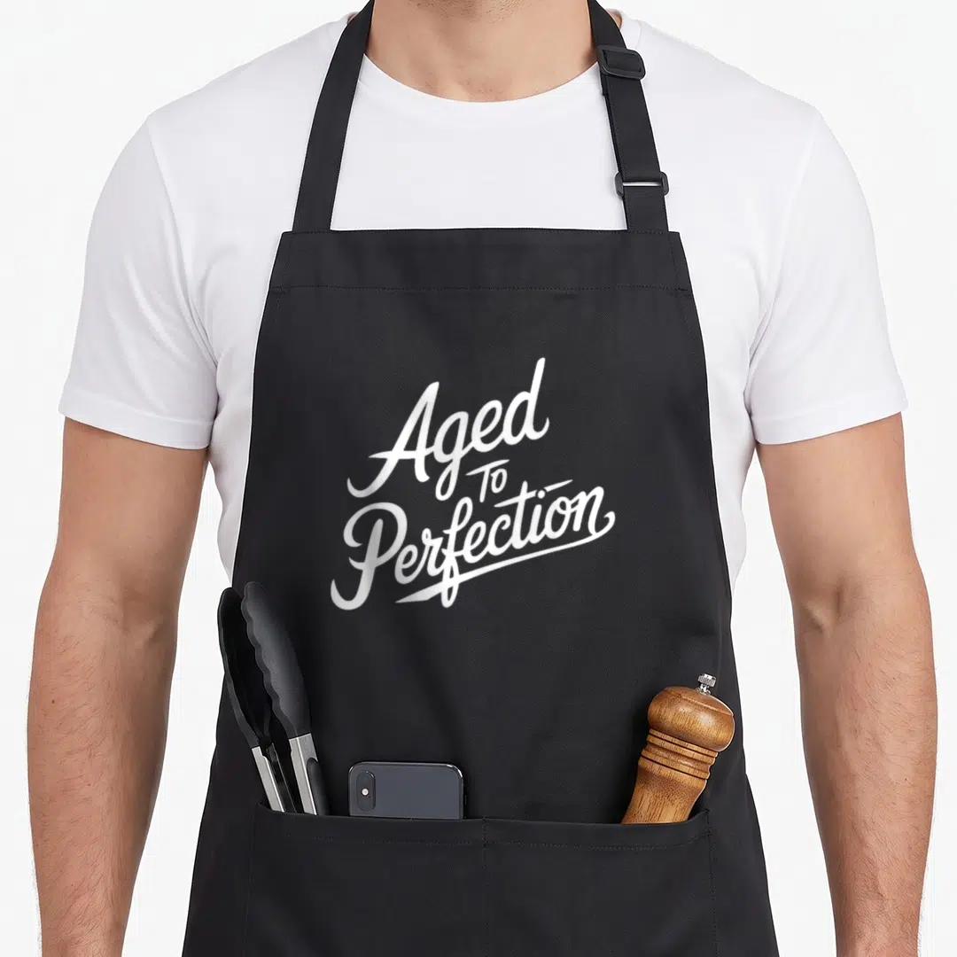 Aged perfectionist apron - White stacked script text reading Aged To Perfection on grey background — shown on a black cotton canvas apron against a white background