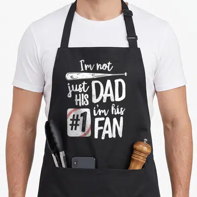 His baseball apron - Distressed white text I'm not just his dad i'm his #1 fan with bat and red stitched baseball — shown on a black cotton canvas apron against a white background