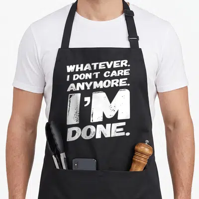 Whatever dont care apron - White stacked text reading WHATEVER I DON'T CARE ANYMORE I'M DONE — shown on a black cotton canvas apron against a white background