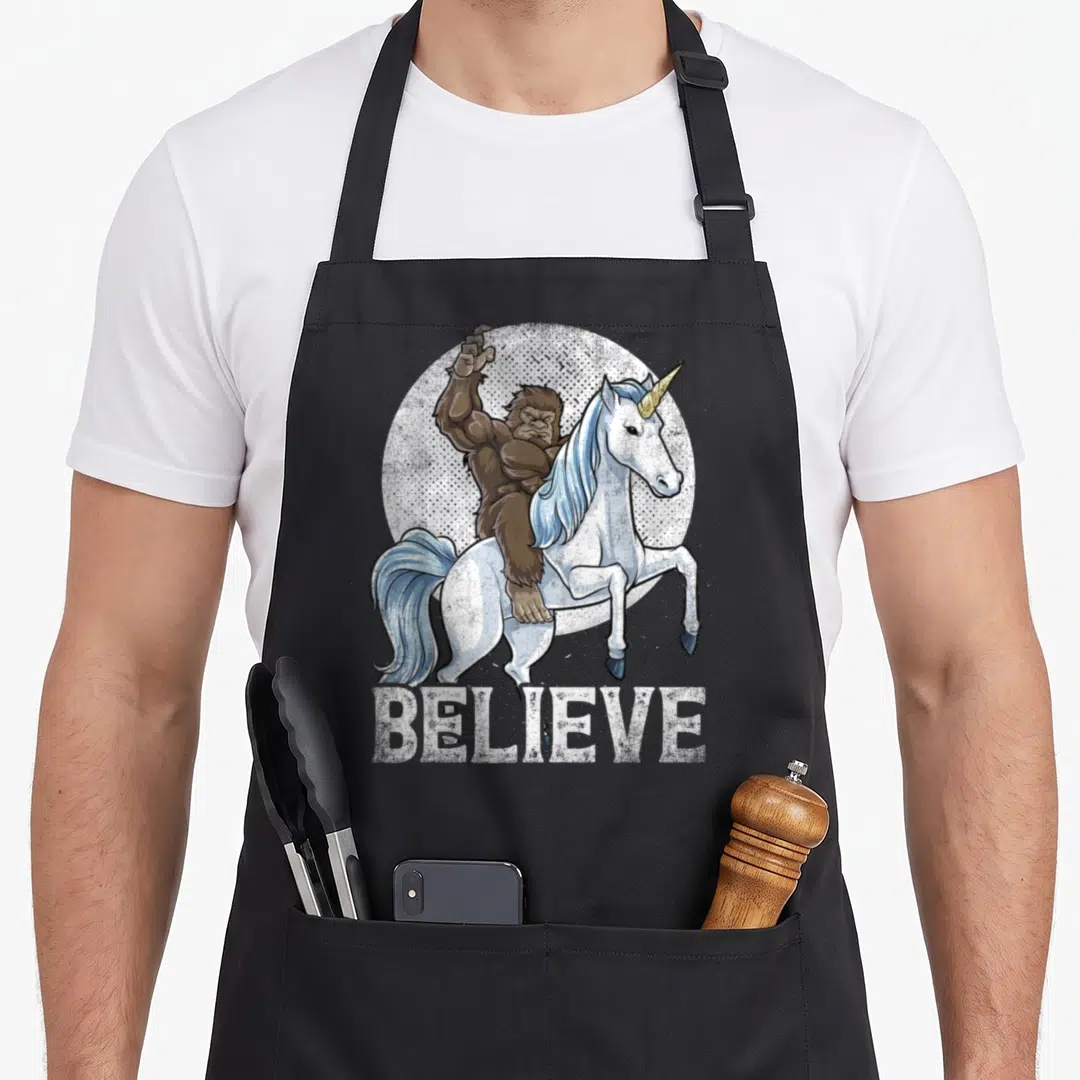 Unicorn believe bigfoot apron - Brown Bigfoot riding white unicorn with blue mane above distressed BELIEVE text — shown on a black cotton canvas apron against a white background