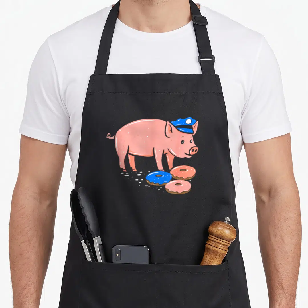 Pig police apron - Cartoon pink pig in blue hat above three pink and blue frosted donuts — shown on a black cotton canvas apron against a white background