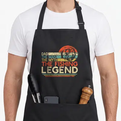 Myth fishing aprons - Dad The Man The Myth The Fishing Legend text with jumping fish in retro striped sun — shown on a black cotton canvas apron against a white background