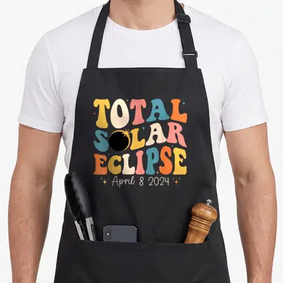 Solar funny event apron - Stacked TOTAL SOLAR ECLIPSE text with eclipsed O and April 8 2024 script — shown on a black cotton canvas apron against a white background