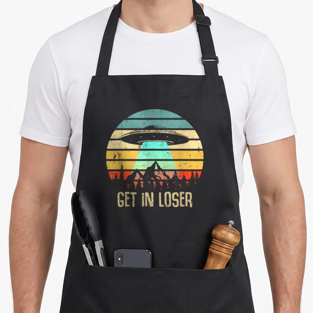 Abduction get loser apron - Retro UFO hovering over mountains with striped sun and GET IN LOSER text — shown on a black cotton canvas apron against a white background