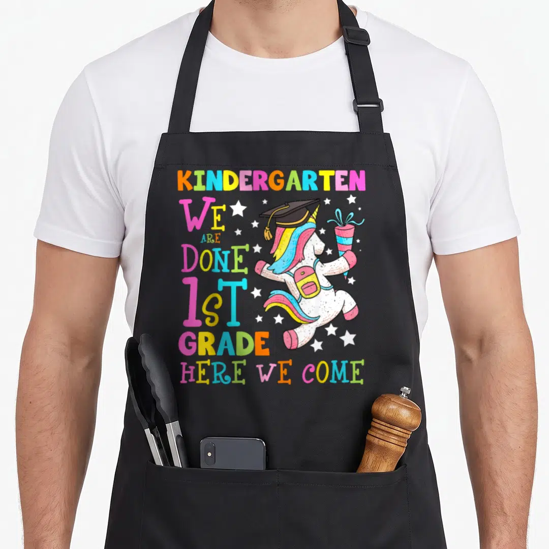 Magical unicorn aprons - Cartoon unicorn with cap backpack and diploma beside stacked colorful text We Are Done 1st — shown on a black cotton canvas apron against a white background