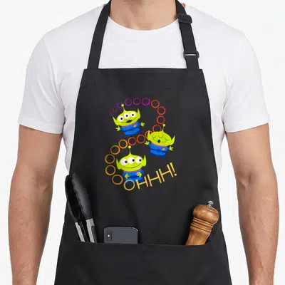 Alien rainbow apron - Three green cartoon aliens in diagonal line with colorful circle trail and OHHH text — shown on a black cotton canvas apron against a white background