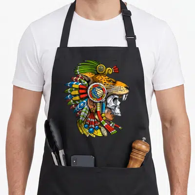 Aztec skull apron - Cartoon jaguar skull human composite head with colorful feathered headdress and gold jewelry — shown on a black cotton canvas apron against a white background