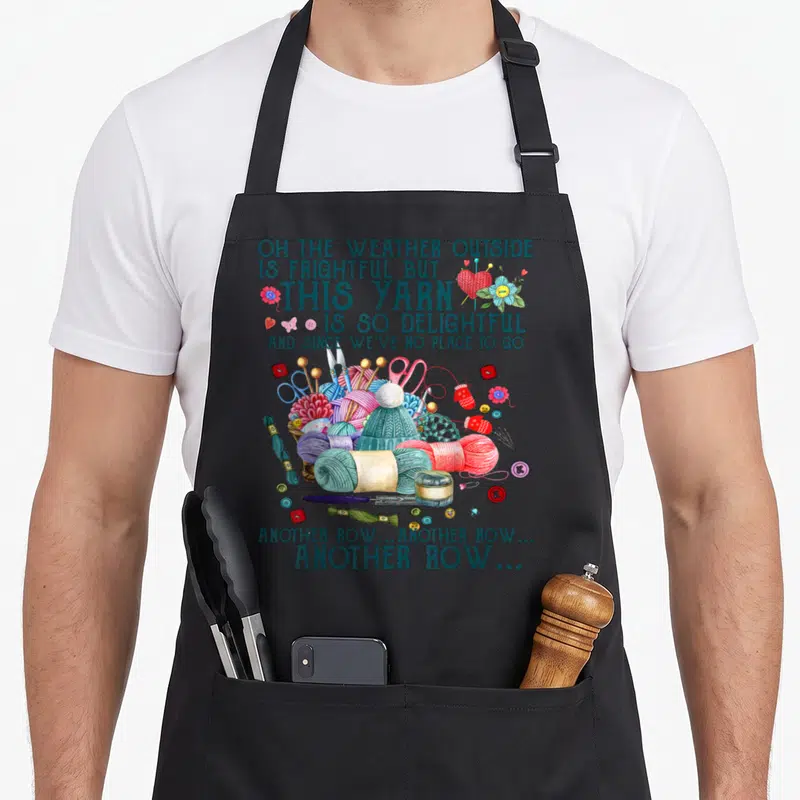Another knitting apron - Cartoon cluster of yarn, scissors, and needles with stacked teal lettering about knitting rows — shown on a black cotton canvas apron against a white background