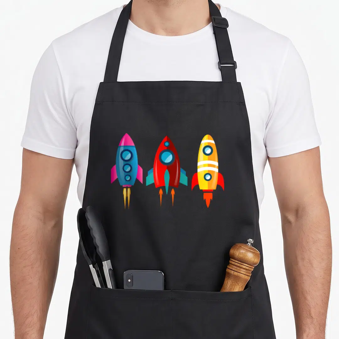 Kosmo cosmo apron - Three cartoon rockets in blue, red, yellow, and orange launching upwards with fire — shown on a black cotton canvas apron against a white background