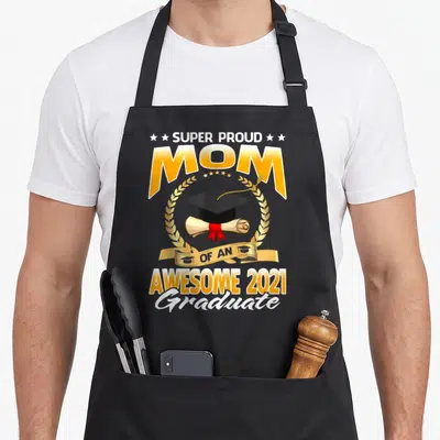 Funny proud graduate apron - Super Proud Mom Of An Awesome 2021 Graduate text with cap and diploma in gold wreath — shown on a black cotton canvas apron against a white background