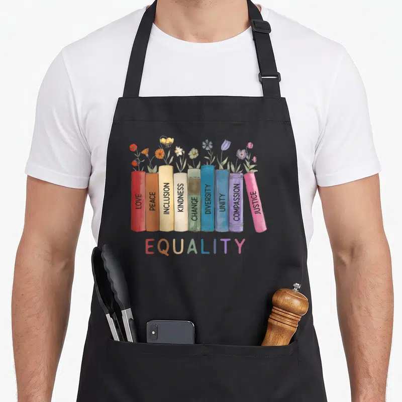 Book floral apron - Nine colorful books with flowers and words like LOVE, PEACE, and EQUALITY — shown on a black cotton canvas apron against a white background