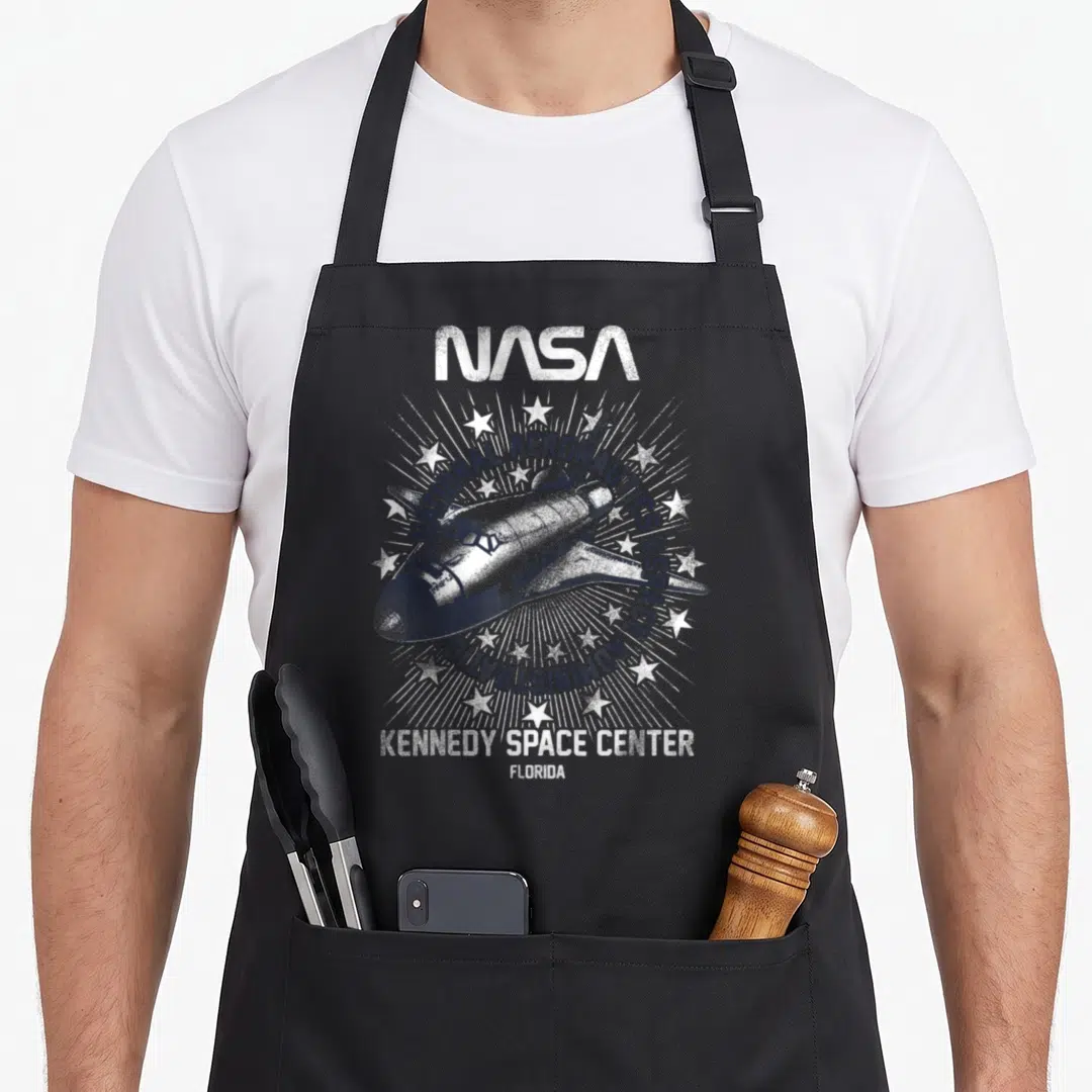 Rocket nasa apron - Distressed white space shuttle surrounded by NASA text and stars on black background — shown on a black cotton canvas apron against a white background