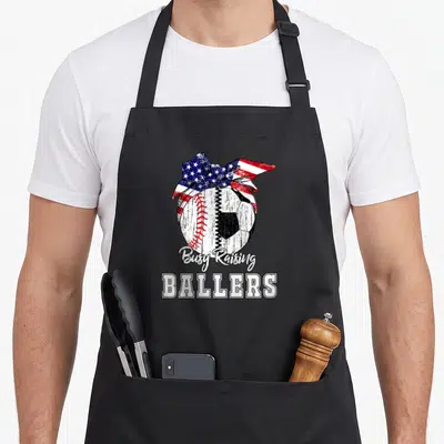 Baseball busy apron - Distressed composite ball with patriotic bow and Busy Raising Ballers text — shown on a black cotton canvas apron against a white background