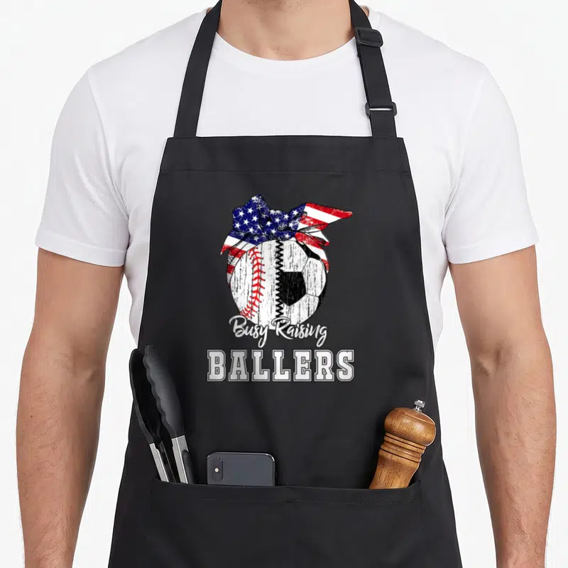 Baseball busy apron - Distressed composite ball with patriotic bow and Busy Raising Ballers text — shown on a black cotton canvas apron against a white background