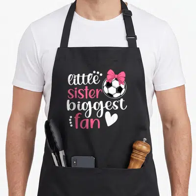 Sister soccer outfit apron - little sister biggest fan text next to a soccer ball with a pink bow and white heart — shown on a black cotton canvas apron against a white background