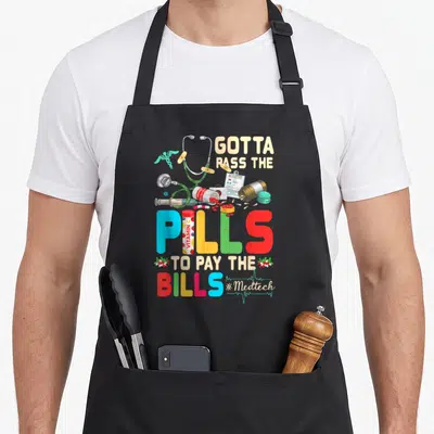 Pass stethoscope and apron - Colorful cartoon pills, syringe, stethoscope, and text Gotta Pass The Pills To Pay — shown on a black cotton canvas apron against a white background