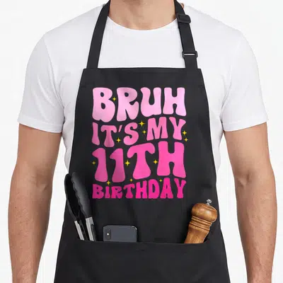 11th 11yr birthday apron - Pink bubble text BRUH IT'S MY 11TH BIRTHDAY with small yellow stars scattered on black fabric — shown on a black cotton canvas apron against a white background