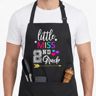 Miss grade second 2nd apron - Stacked text little MISS 2ND Grade with pencil I, hearts, and yellow arrow in pink blue — shown on a black cotton canvas apron against a white background