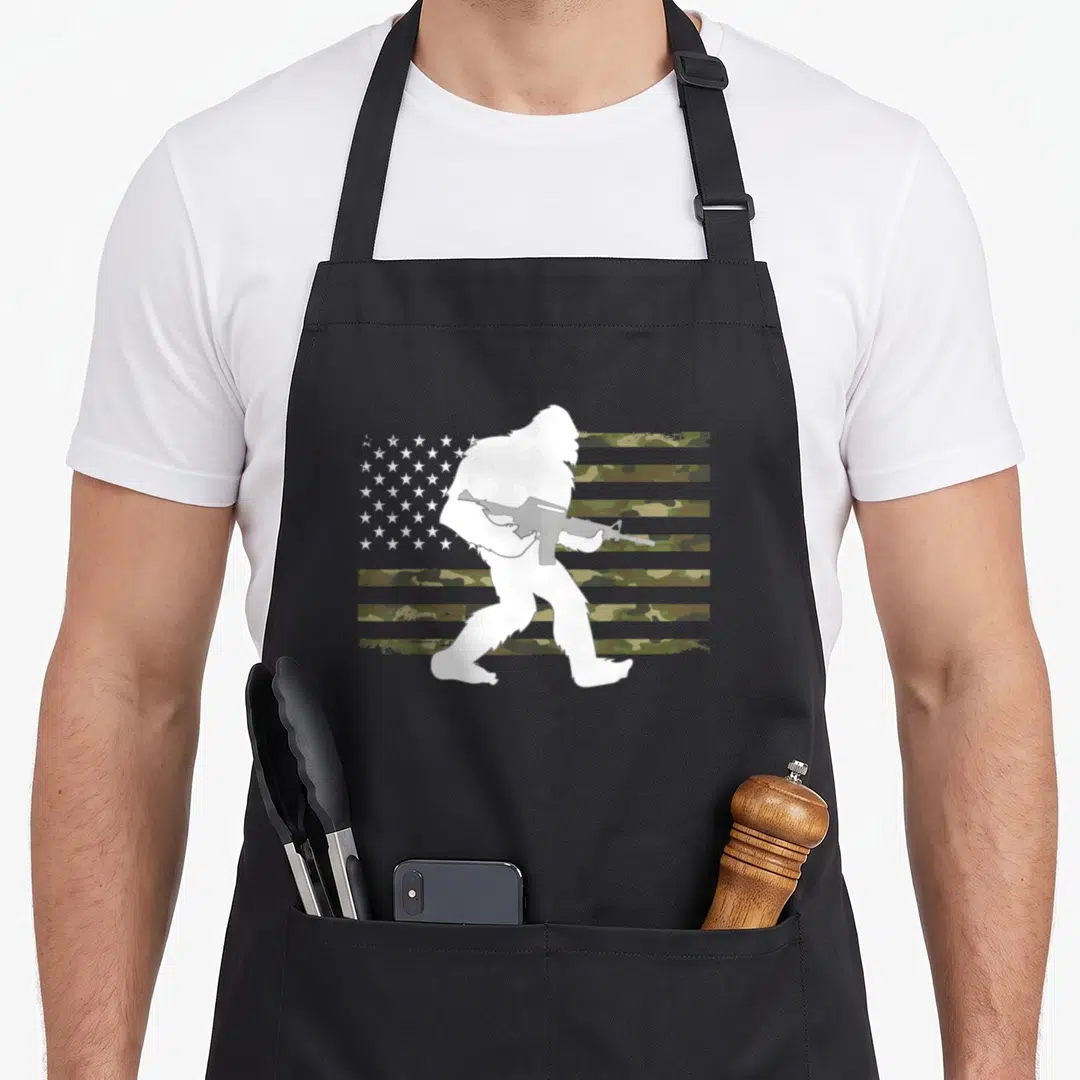Bigfoot camo aprons - White Bigfoot silhouette holding rifle in front of green brown camo US flag stripes — shown on a black cotton canvas apron against a white background
