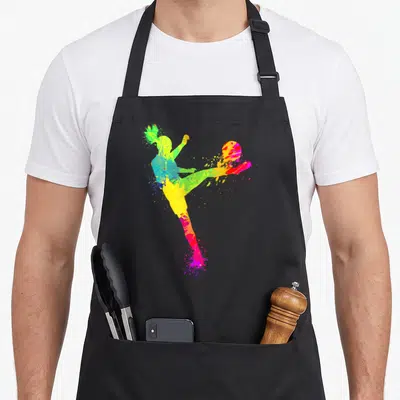 Player funny soccer apron - Multi-color paint splatter silhouette of a soccer player kicking a ball in yellow magenta — shown on a black cotton canvas apron against a white background