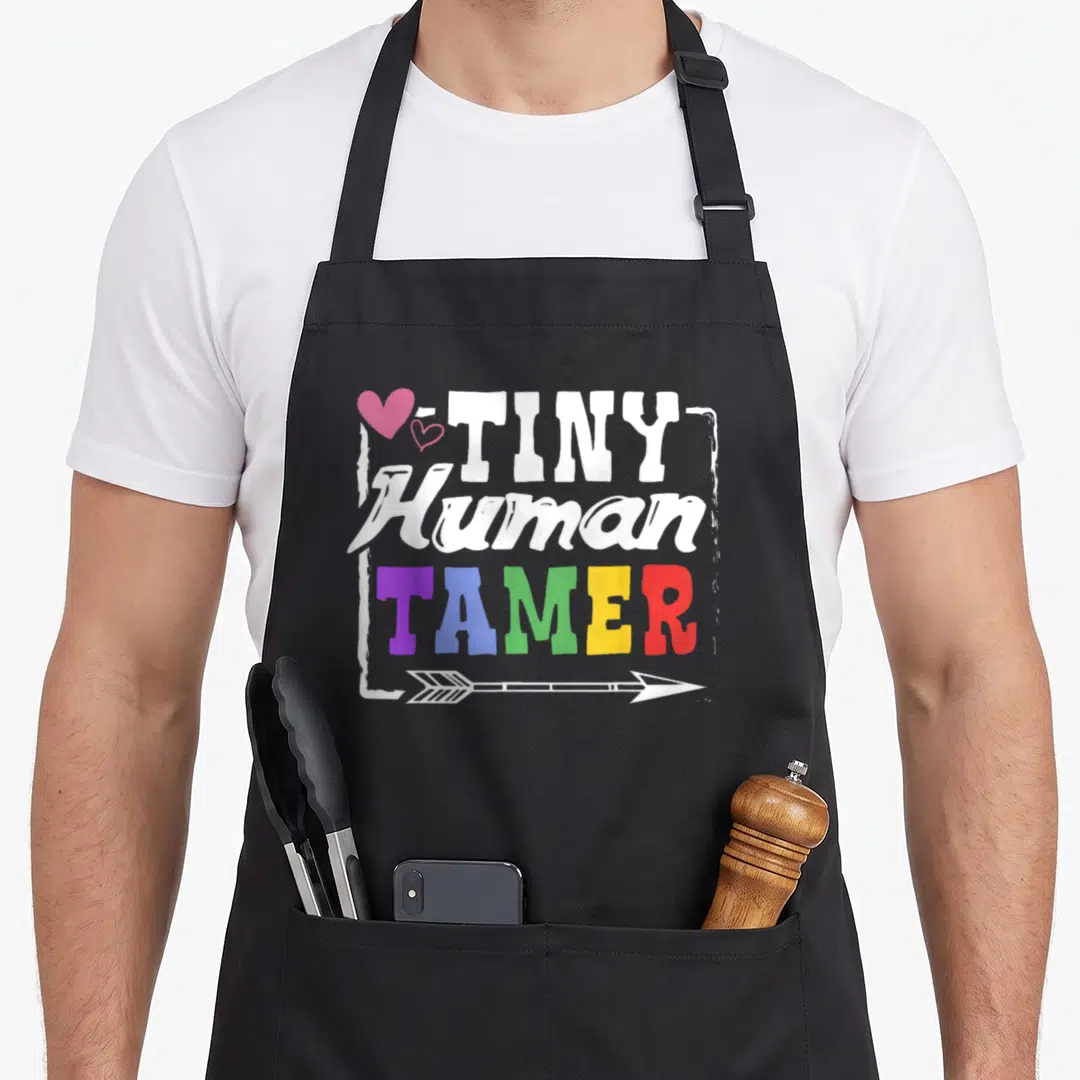 Teacher tiny chef apron - Stacked TINY Human TAMER text with pink hearts and arrow in rainbow colors — shown on a black cotton canvas apron against a white background