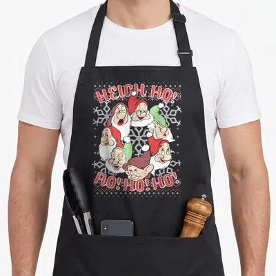 Snow ugly christmas sweater apron - Six cartoon dwarf heads with arched HEIGH HO text and snowflakes in red green white — shown on a black cotton canvas apron against a white background