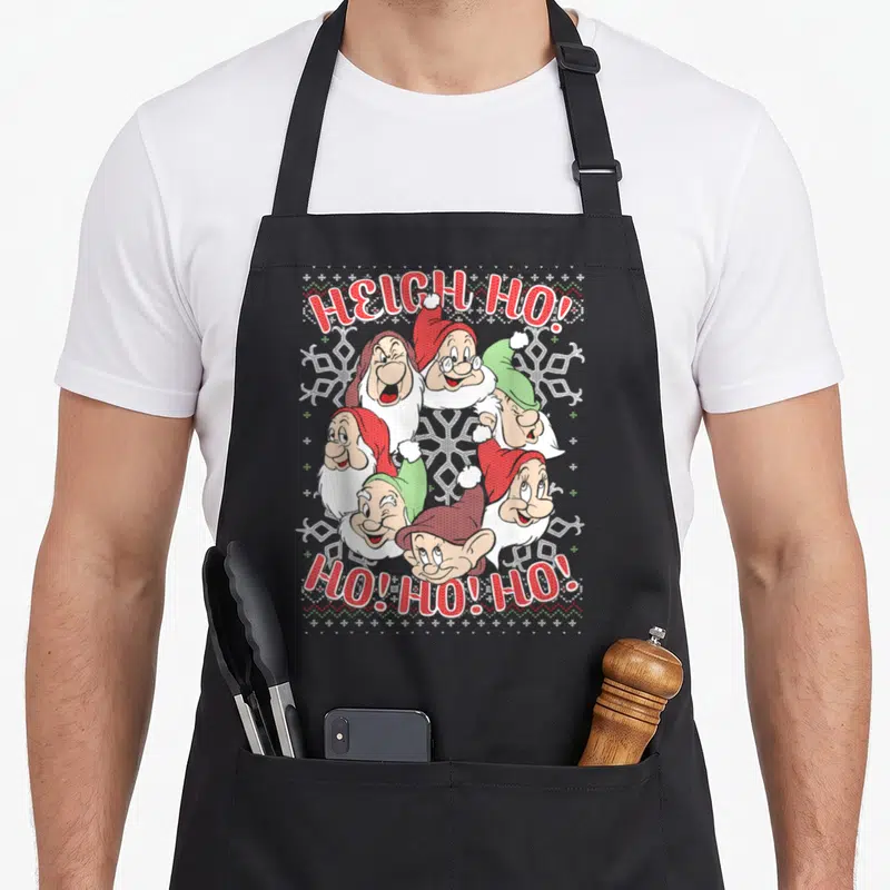 Snow ugly christmas sweater apron - Six cartoon dwarf heads with arched HEIGH HO text and snowflakes in red green white — shown on a black cotton canvas apron against a white background