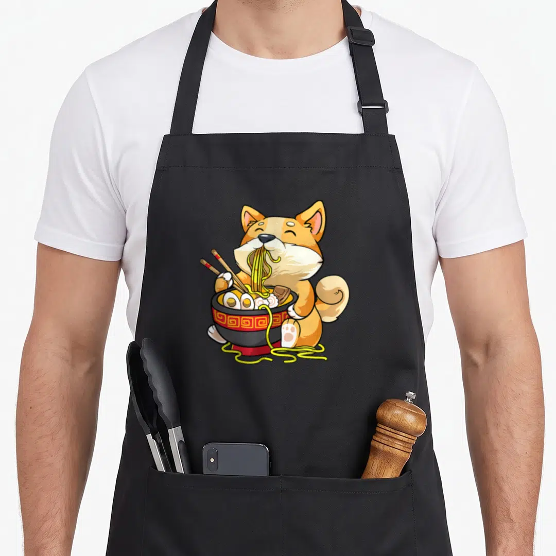 Corgi pho apron - Cartoon dog eating yellow noodles from red and black bowl with chopsticks — shown on a black cotton canvas apron against a white background