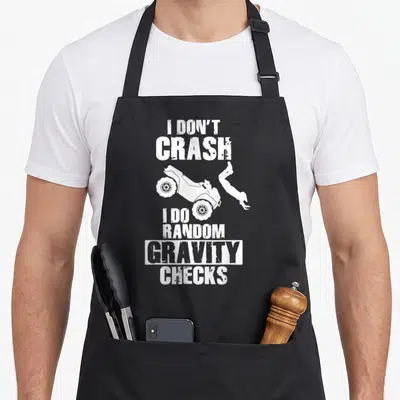 Crash vehicle apron - White text I Don't Crash I Do Random Gravity Checks above and below ATV rider silhouette — shown on a black cotton canvas apron against a white background