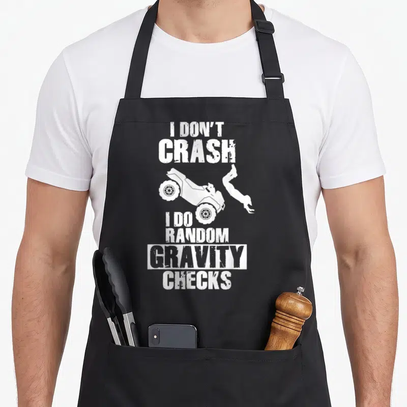 Crash vehicle apron - White text I Don't Crash I Do Random Gravity Checks above and below ATV rider silhouette — shown on a black cotton canvas apron against a white background