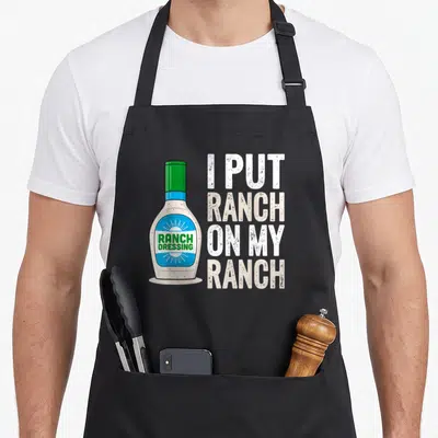 Dressing ranch vibes apron - Cartoon ranch dressing bottle next to stacked text reading I put ranch on my ranch — shown on a black cotton canvas apron against a white background