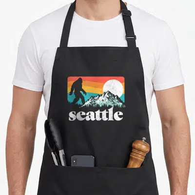 Bigfoot seattle apron - Retro graphic with Bigfoot silhouette, striped background, mountains, white sun, and seattle — shown on a black cotton canvas apron against a white background