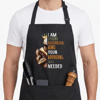 Horoscope king apron - Crowned man illustration beside stacked text I AM STRONG AQUARIUS KING YOUR APPROVAL Isn't — shown on a black cotton canvas apron against a white background