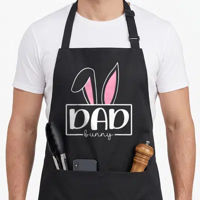 Family easter bunny aprons for women - DAD bunny text with two pink ears emerging from white rectangular frame — shown on a black cotton canvas apron against a white background
