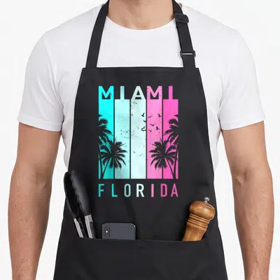 Miami florida apron - Retro MIAMI FLORIDA text over cyan white pink stripes with palm tree and bird silhouettes — shown on a black cotton canvas apron against a white background