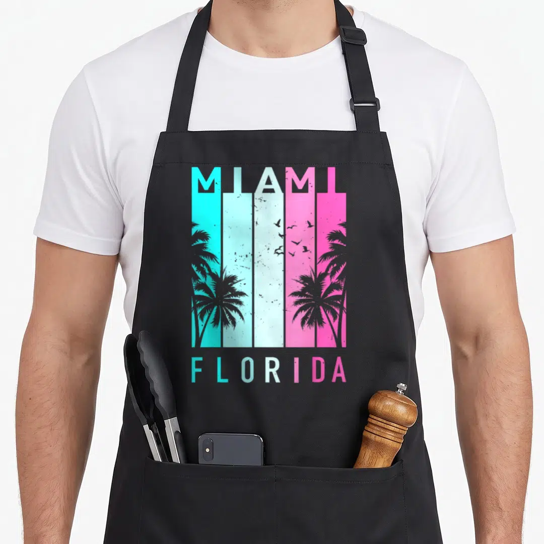 Miami florida apron - Retro MIAMI FLORIDA text over cyan white pink stripes with palm tree and bird silhouettes — shown on a black cotton canvas apron against a white background