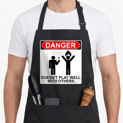 Danger antisocial apron - Warning sign with red DANGER oval, two black stick figures, one holding a lit match near — shown on a black cotton canvas apron against a white background