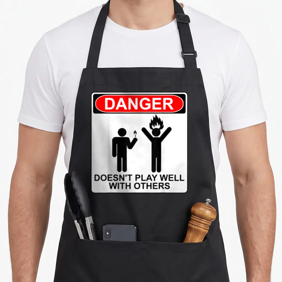 Danger antisocial apron - Warning sign with red DANGER oval, two black stick figures, one holding a lit match near — shown on a black cotton canvas apron against a white background