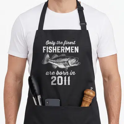 15th fishing aprons - Distressed white fish illustration between text blocks reading Only the finest FISHERMEN are born — shown on a black cotton canvas apron against a white background