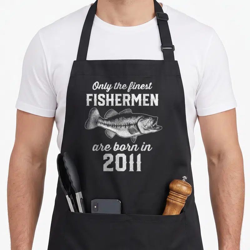15th fishing aprons - Distressed white fish illustration between text blocks reading Only the finest FISHERMEN are born — shown on a black cotton canvas apron against a white background