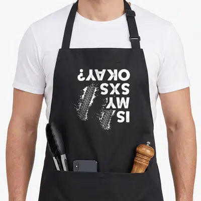 Sxs utv girl apron - White rotated text IS MY SXS OKAY? beside two distressed tire tracks with mud splashes — shown on a black cotton canvas apron against a white background