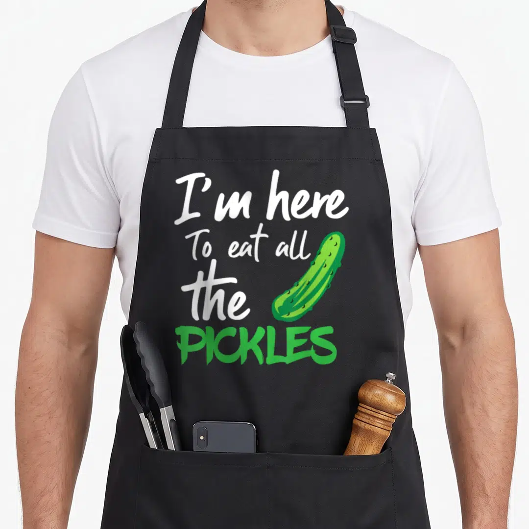 Cute eat pickle apron - Stacked white and green text reading I'm here To eat all the PICKLES beside a green cartoon — shown on a black cotton canvas apron against a white background
