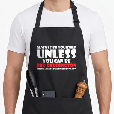 Alway redding apron - Stacked white and red text reading Always Be Yourself Unless You Can Be Red Reddington — shown on a black cotton canvas apron against a white background