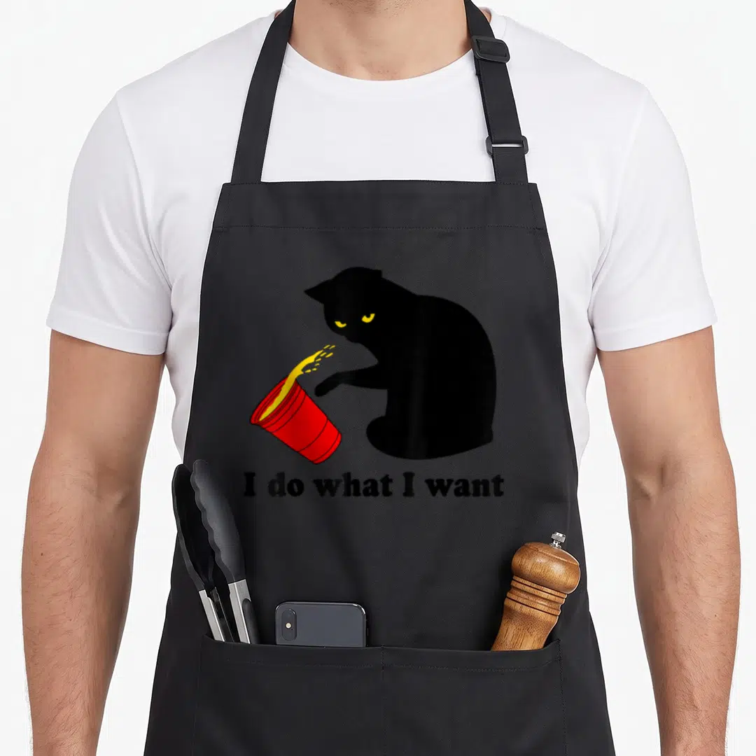 Cat dark red apron - Black cat tipping red cup with yellow liquid and text I do what I want — shown on a black cotton canvas apron against a white background