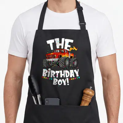 Son truck aprons - Red monster truck with THE BIRTHDAY BOY! text and confetti — shown on a black cotton canvas apron against a white background