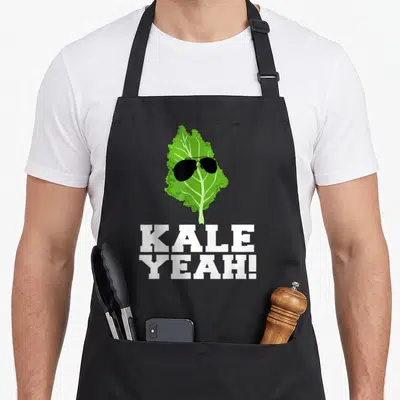Yeah vegan apron - Green kale leaf wearing black sunglasses above stacked white KALE YEAH! text — shown on a black cotton canvas apron against a white background