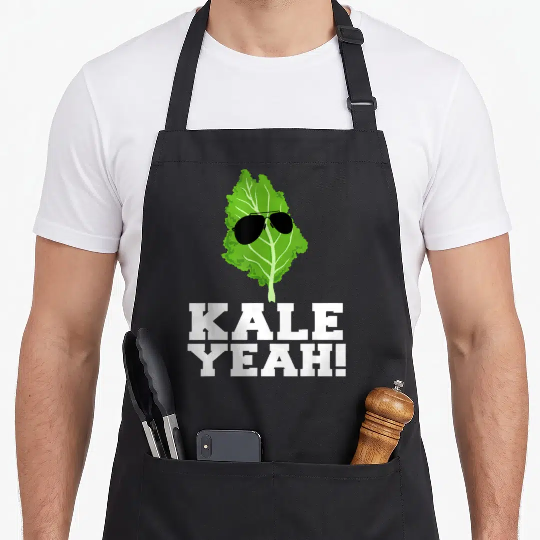 Yeah vegan apron - Green kale leaf wearing black sunglasses above stacked white KALE YEAH! text — shown on a black cotton canvas apron against a white background