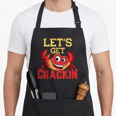 Crackin crabbing crab apron - Cartoon red crab between yellow LET'S GET and red CRACKIN text — shown on a black cotton canvas apron against a white background